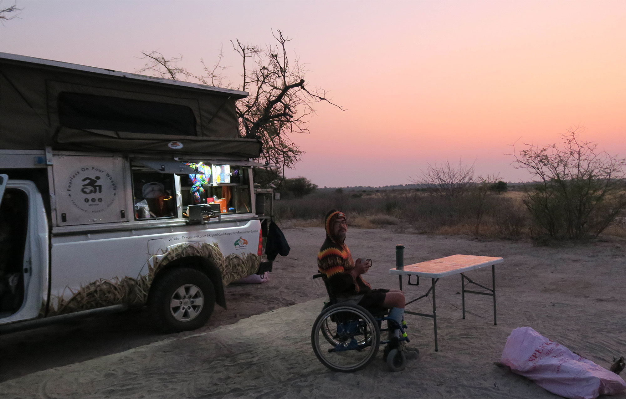 Man is in a wheel chair in an epic road trip voyage. He is seen at sunset in a wheelchair drinking water near his accessible van.