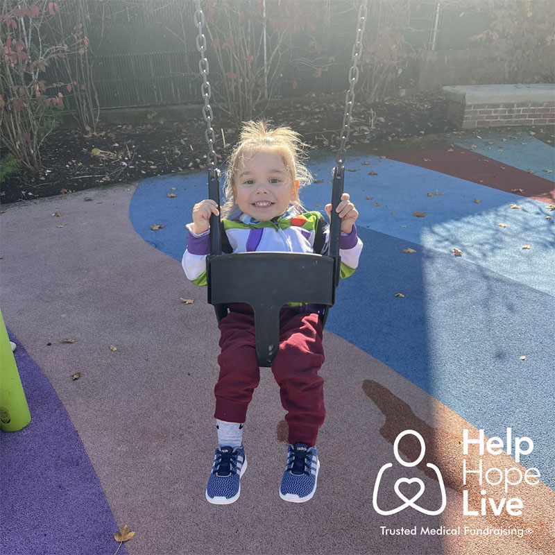 A little boy sits on the playground equipment at the park and smiles. The Help Hope Live logo appears in the lower righthand corner.