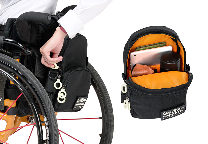 A woman is reaching back to unzip the black FEELDOM JAYU wheelchair bag attached to the back of her wheelchair. Another open bag features an orange interior with glasses case, tablet and thermos.