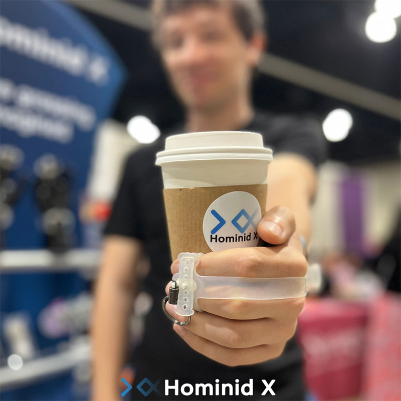 Man with brown hair holding a coffee cup using Fiber, a grasping device designed for those with hand weakness. The coffee cup has the Hominid X logo on it. 