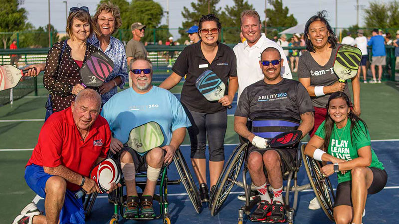A group of pickleball players gather after a match. Two use wheelchairs and the other players are ambulatory.