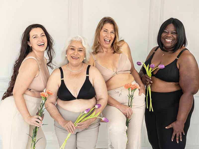 Four smiling women of various ages, sizes and ethnicities are holding flowers and modeling beige and black Springrose adaptive bras.