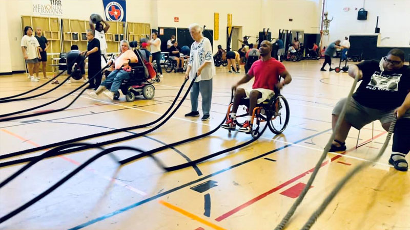 Five people at an Adaptive Tabata Fitness Class. Some use wheelchairs and others are standing. They all hold the ends of heavy ropes used to build arm strength.