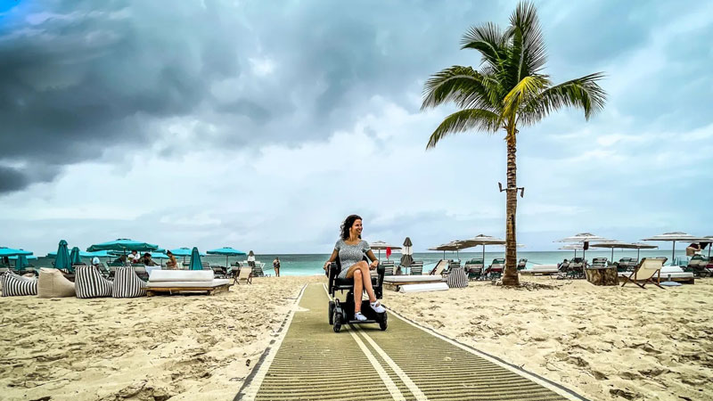 Accessible beach shows a woman who uses a wheelchair riding an accessible pathway over the sand at a beach.