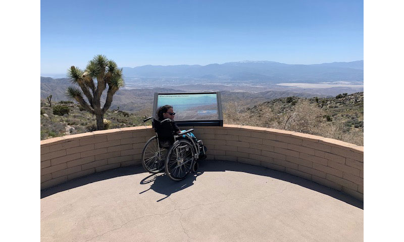A young woman in a wheelchair sits at a lookout point with the picturesque Joshua Tree National Park and the distant mountains in the background.